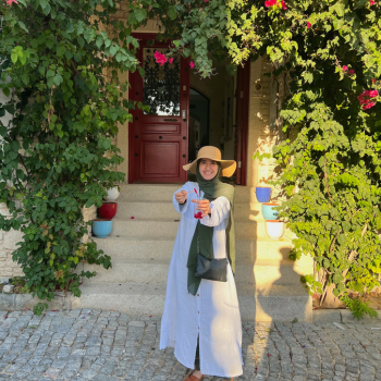 Dr. Zeynap Ozdener smiling in a garden in front of a building with a red door wearing a white dress, green scarf and sun hat
